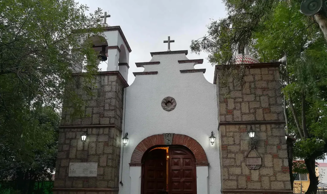 Fachada de la capilla de piedra y muro blanco con campanario y puerta de madera