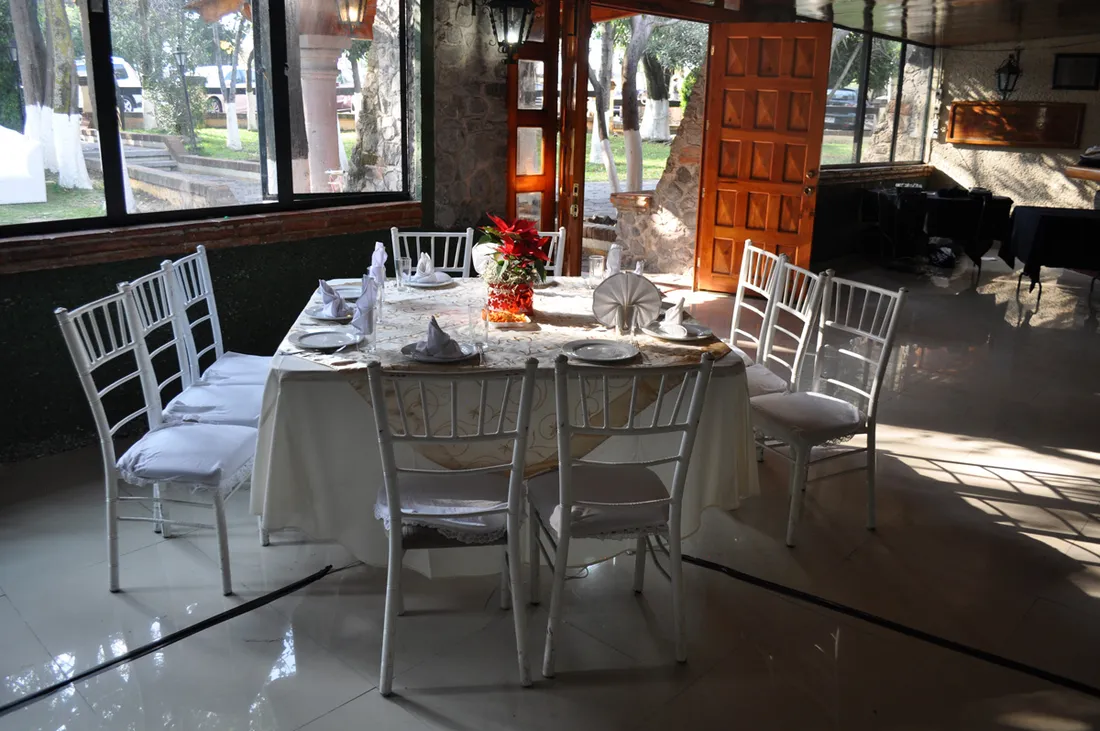 Mesa de cena con sillas blancas junto a ventanales que dan a los jardines